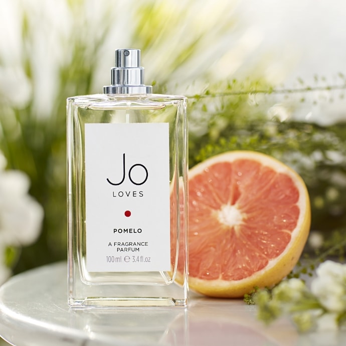 bootle of Jo Love perfume in a see through bottle on a table with a half cut orange in the background.